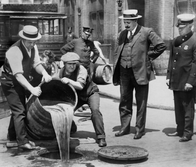 Men pouring alcohol into a street drain while police officials watch