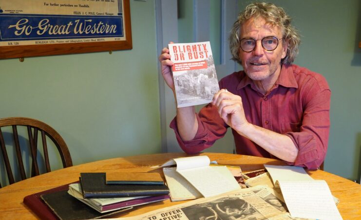 David Wilkins holding up a copy of 'Blighty or Bust' while sitting at his kitchen table