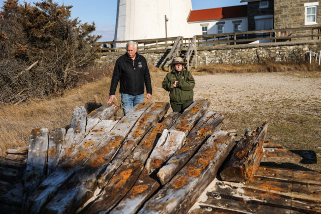 Two people stand behind a large wooden piece of shipwreck on the shores of a beach.